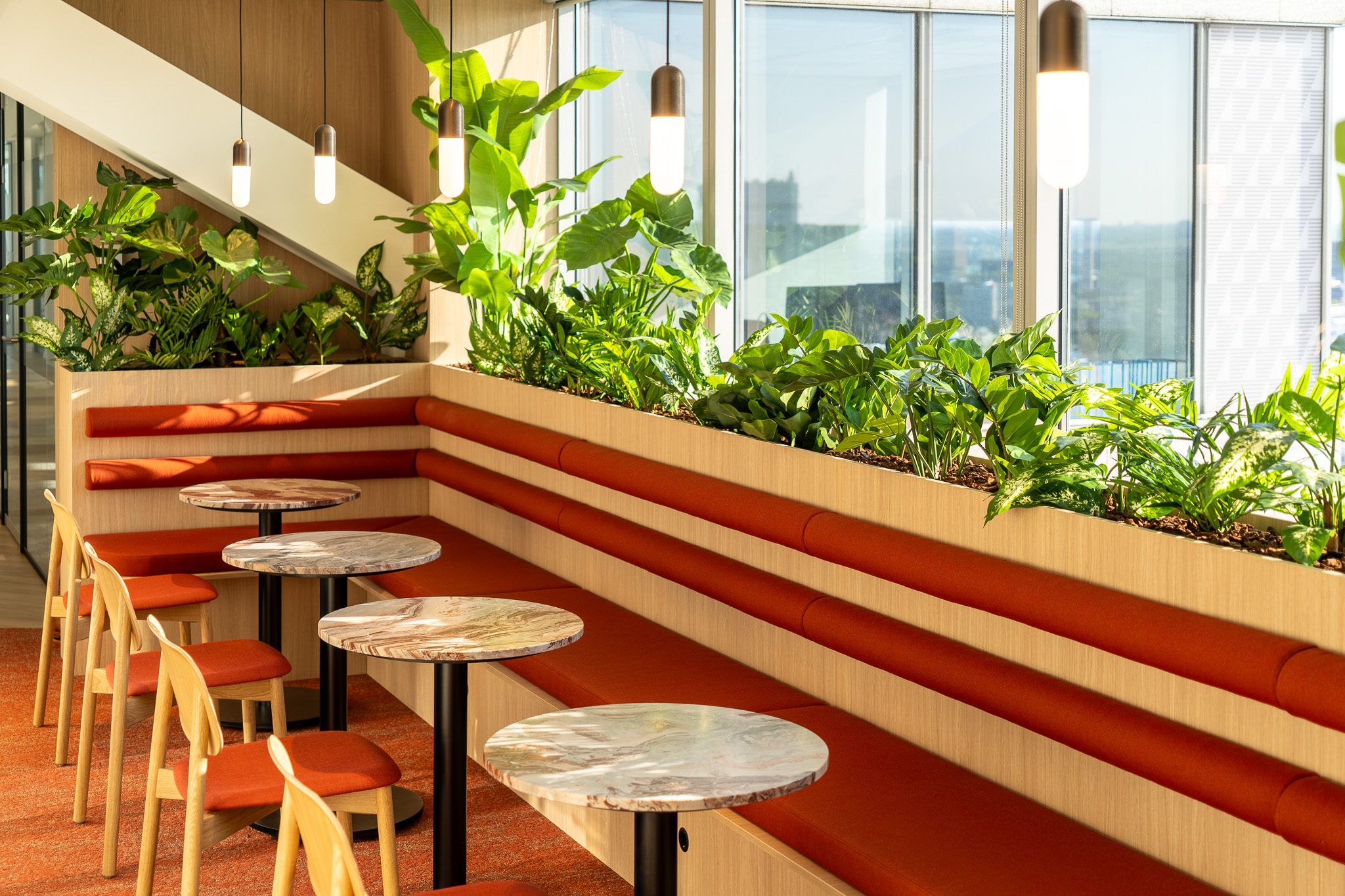 Lange bank met oranje kussens langs een plantenbak vol groen, ronde marmeren tafeltjes en glazen hanglampen voor de ramen van een kantoor aan de Blaak