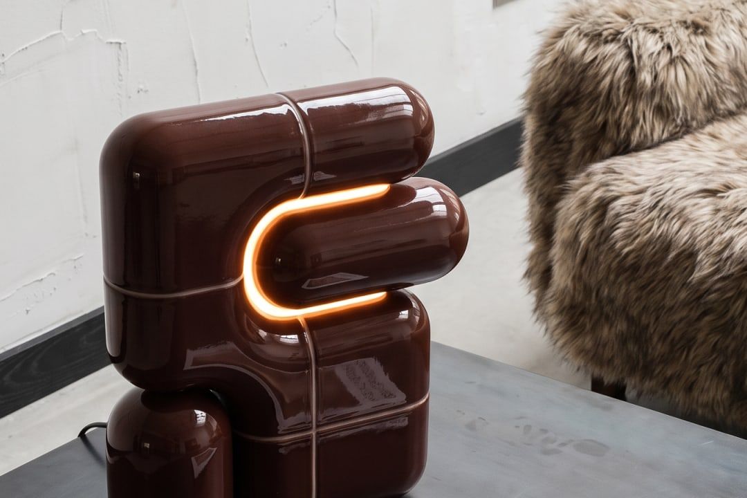 Kleine lamp in hoogglanzend donkerbruin, opgebouwd uit gebogen buisvormen met een oplichtende U-lijn, op een donker tafelblad met een tijdschrift, naast een fauteuil van lang bont