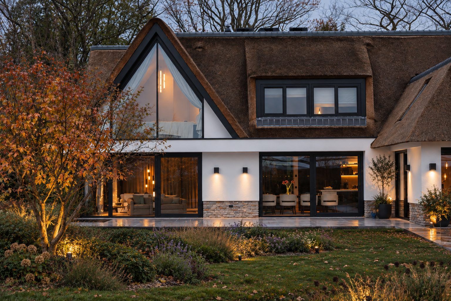 De woning met rieten dak en een glazen puntgevel bij schemering, met licht achter de ramen, wandlampen op de gevel en aangelichte bomen en borders in de tuin