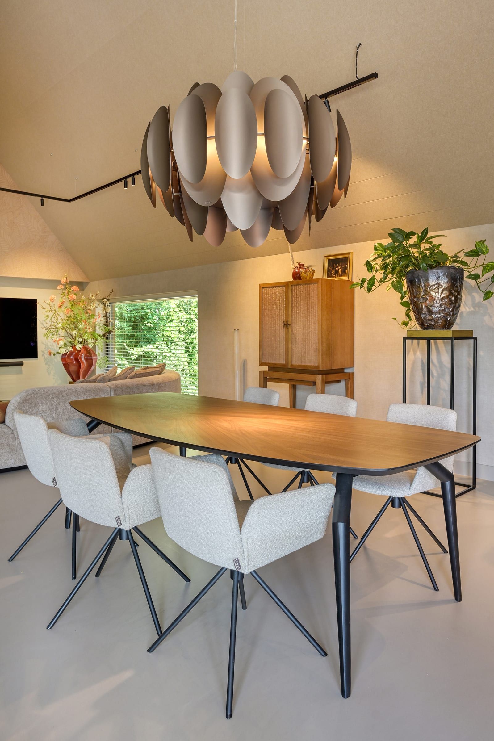Oval timber dining table with pale chairs beneath a large pendant of curved bronze leaves, in a living room with a sloping ceiling and a window onto the garden