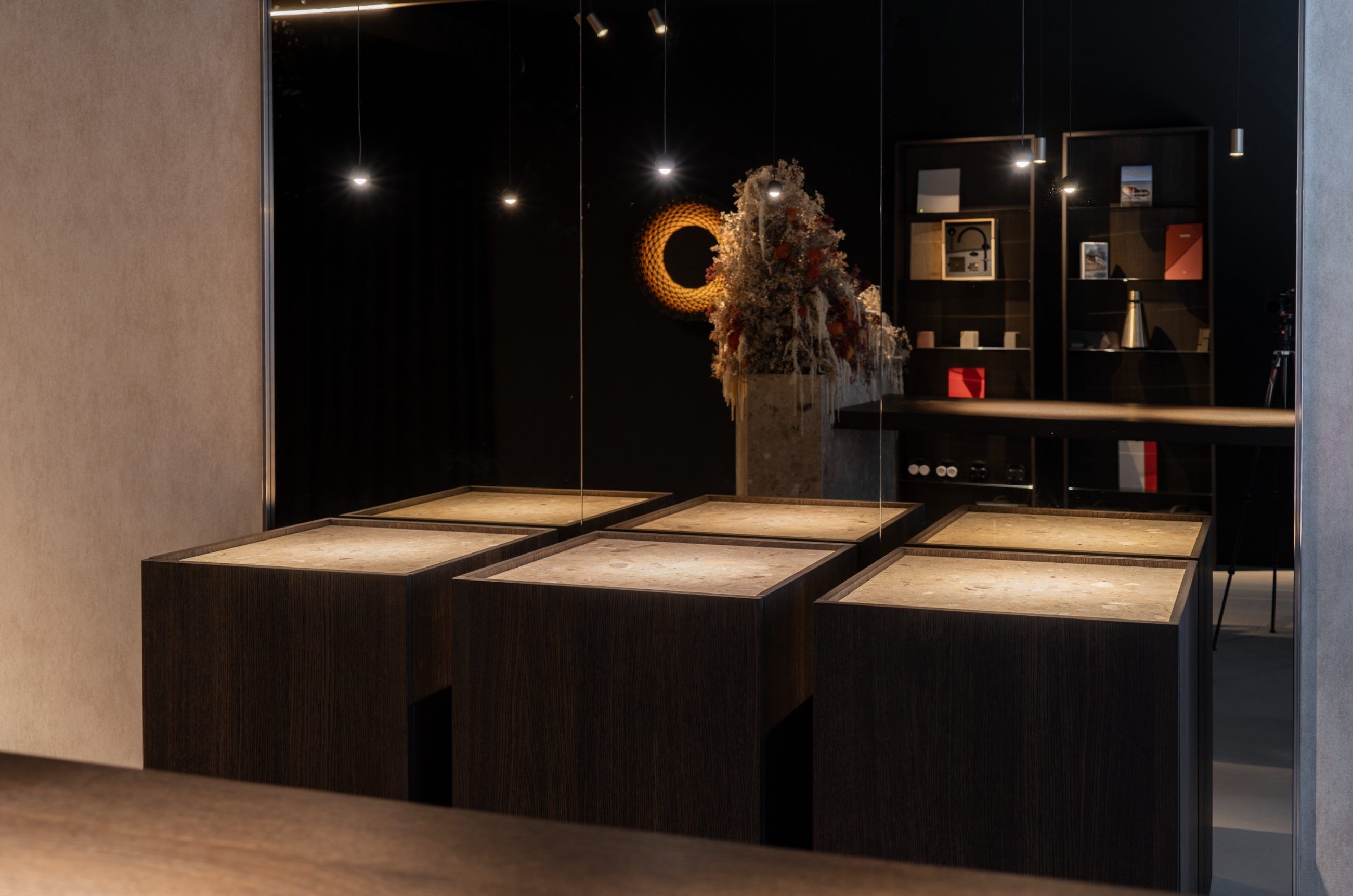 Vier verlichte vitrinetafels van donker hout voor een glazen wand, met een zuil met droogbloemen en een ronde wandlamp erachter