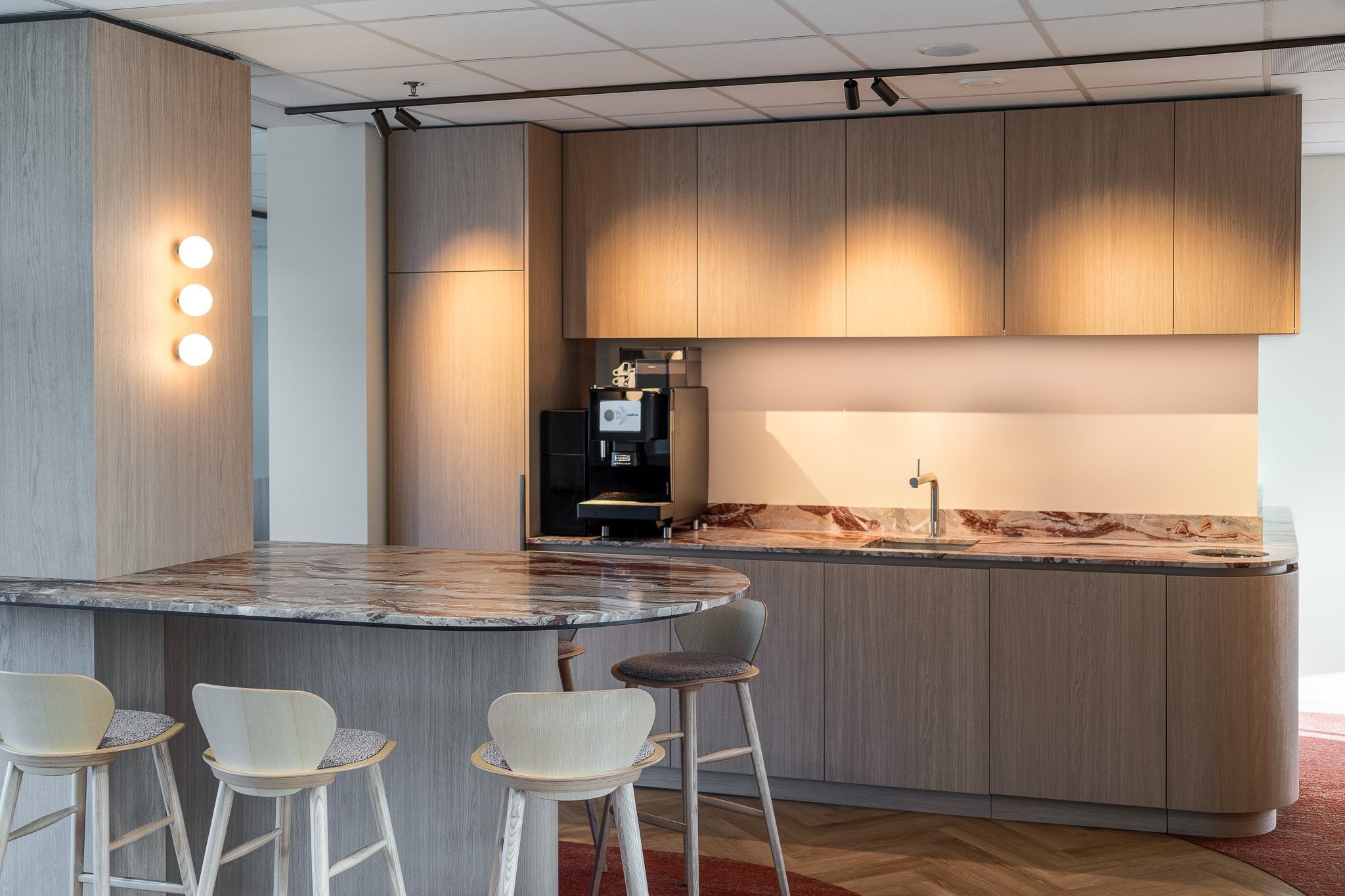 Pantry met houten kastenwand, strijklicht van spots op de fronten, een marmeren bar met houten krukken en drie ronde wandlampen op een zuil