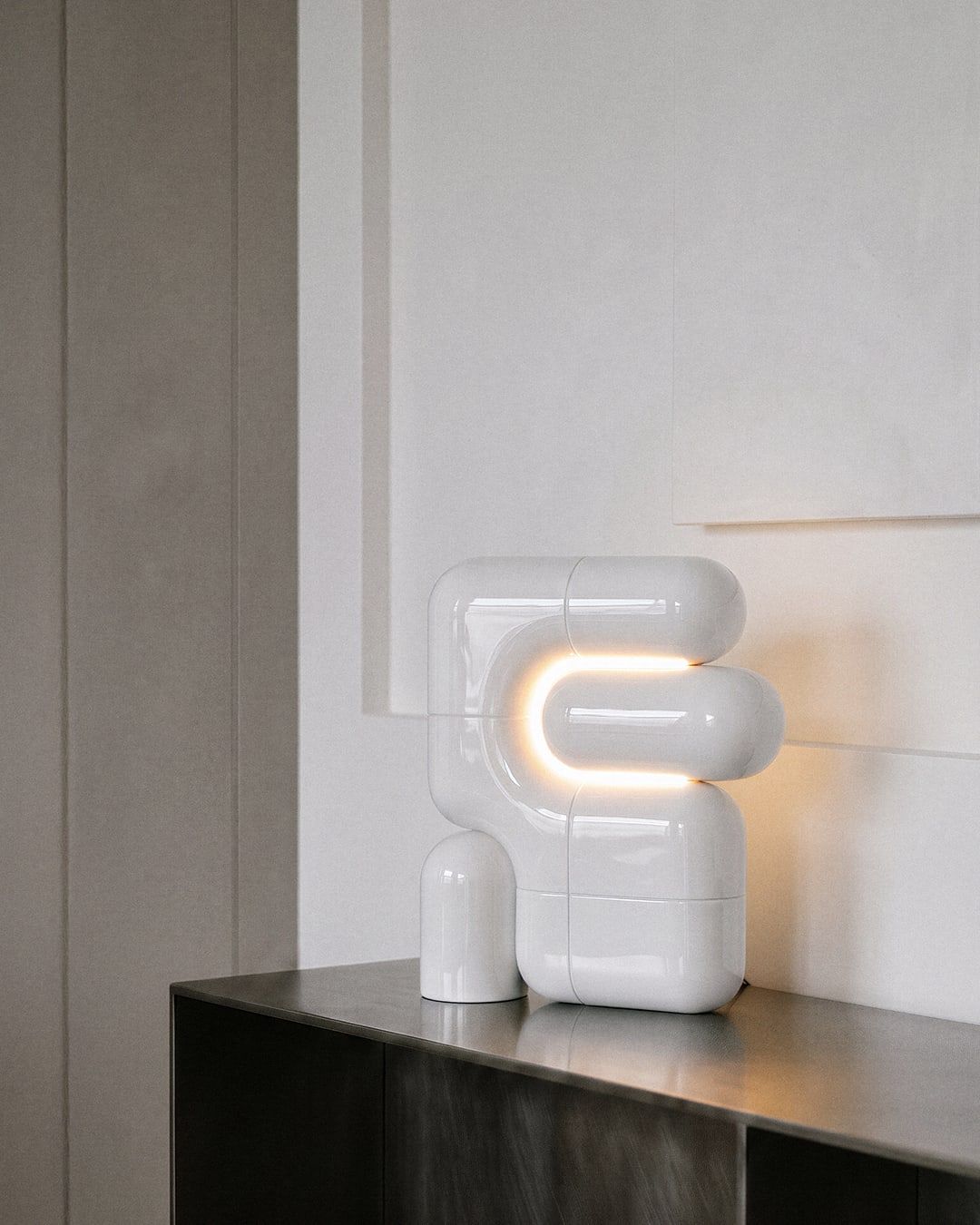 Witte hoogglanzende lamp uit gebogen buisvormen met een oplichtende U-lijn, boven een donker dressoir tegen een lichte wand