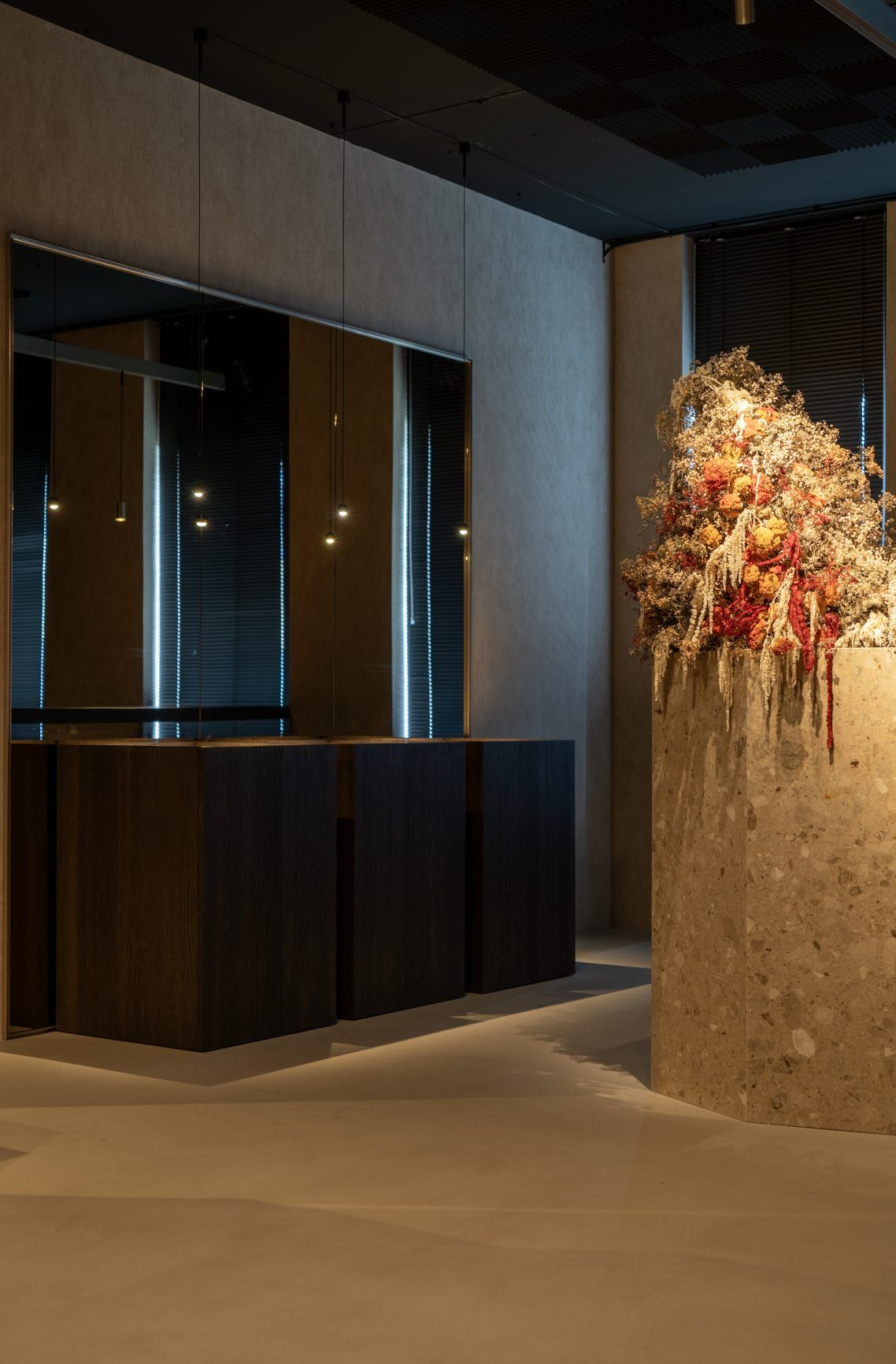 Zuil van terrazzo met een bloemstuk van droogbloemen, in het licht van smalle hanglampen voor een rij vitrines