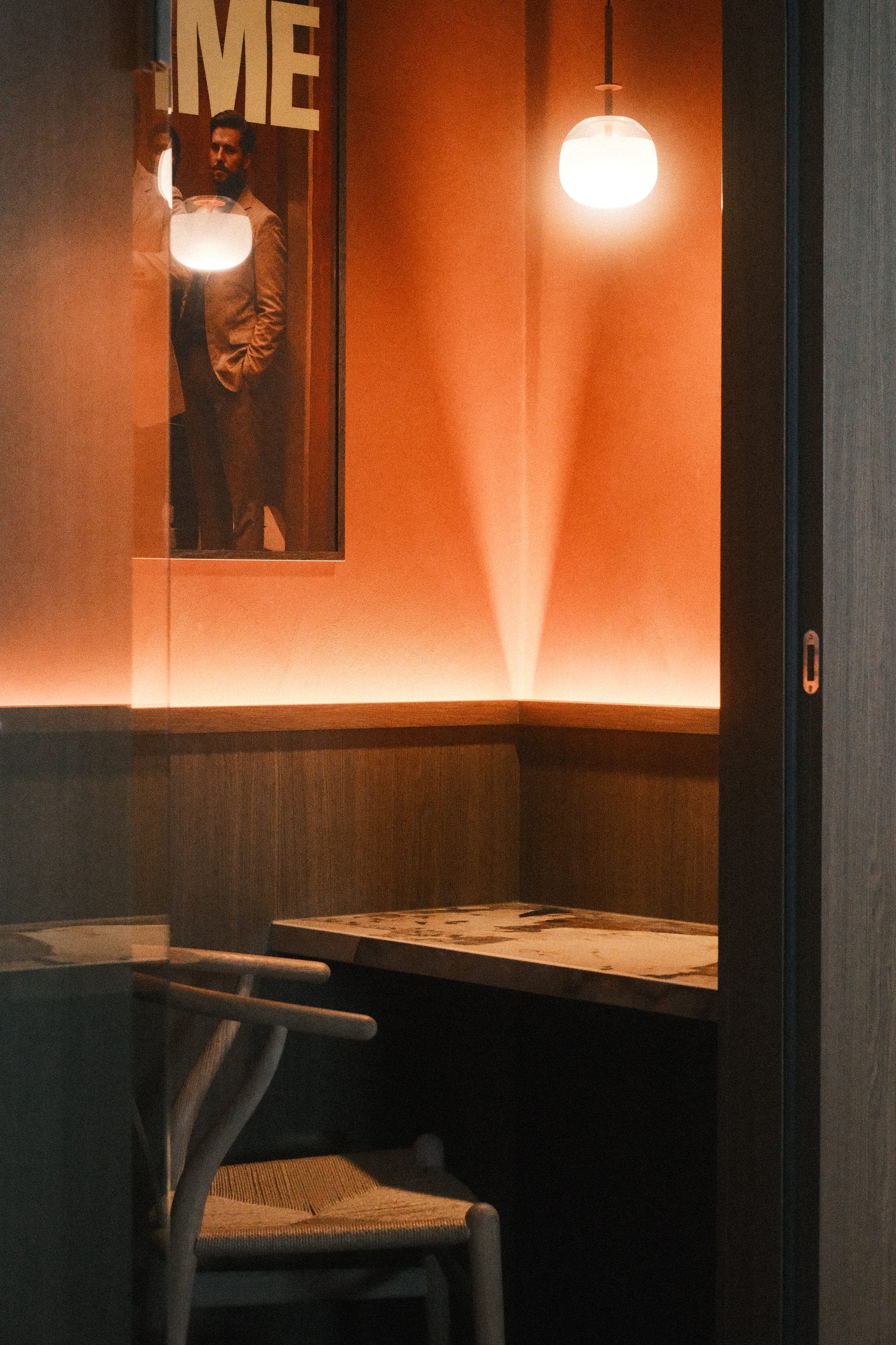 Belcel met een marmeren tafeltje en een stoel, een glazen bol als hanglamp en een warme lichtlijn tegen een oranje wand
