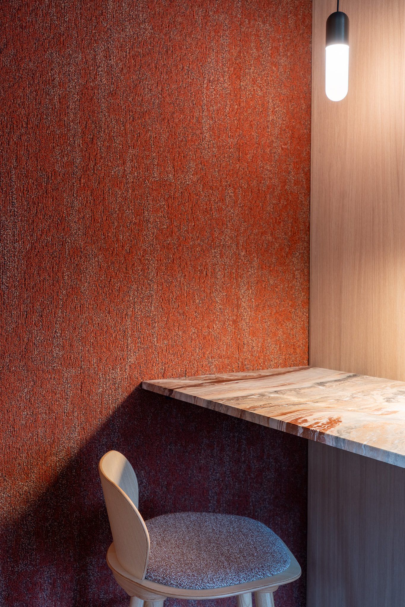 Glazen hanglamp boven een smalle marmeren tafel tegen een wand van roestrood textiel, met een houten stoel