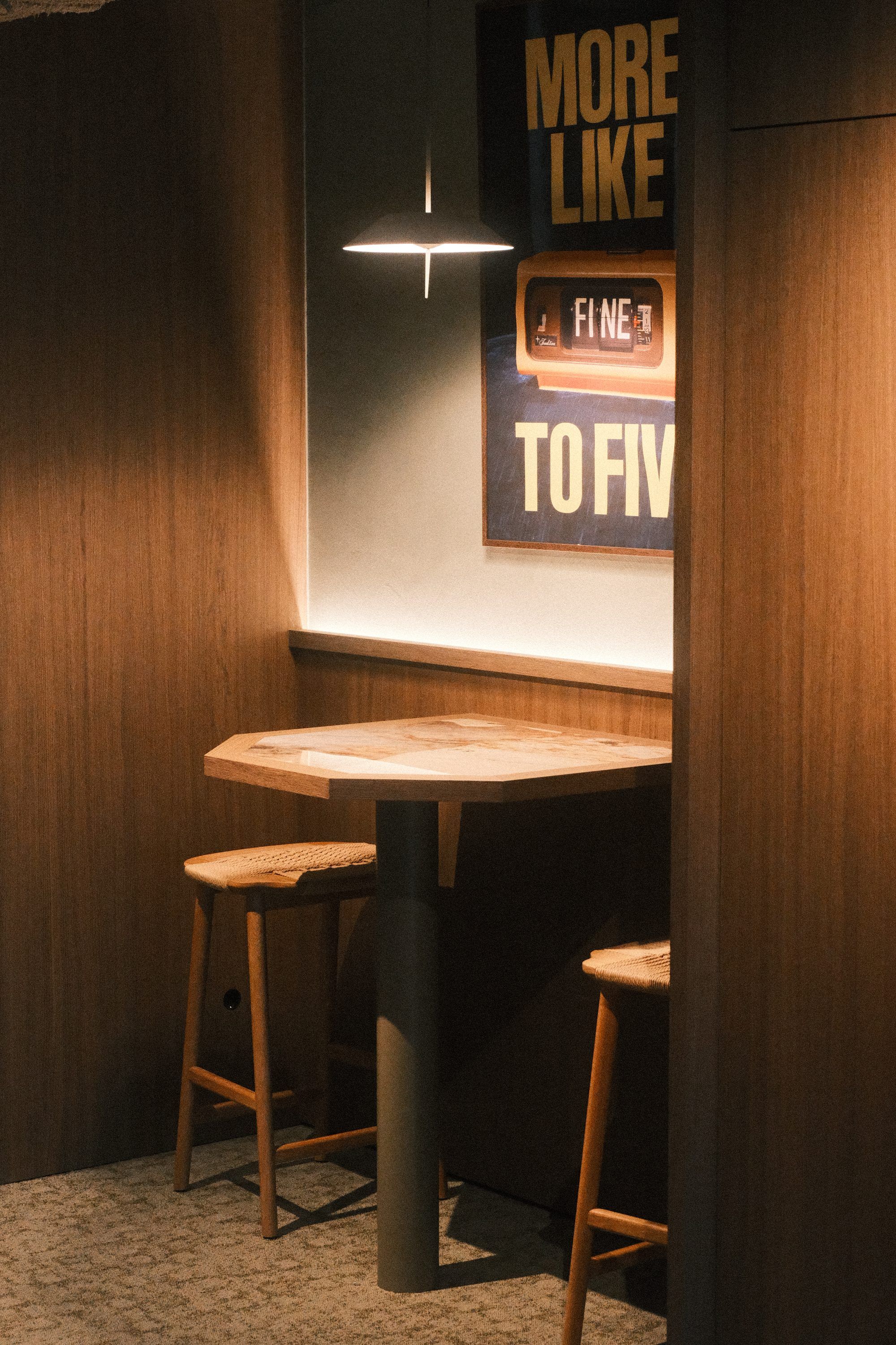 Nis met een statafel en krukken onder een platte hanglamp, met een lichtlijn boven de houten lambrisering en een poster aan de wand