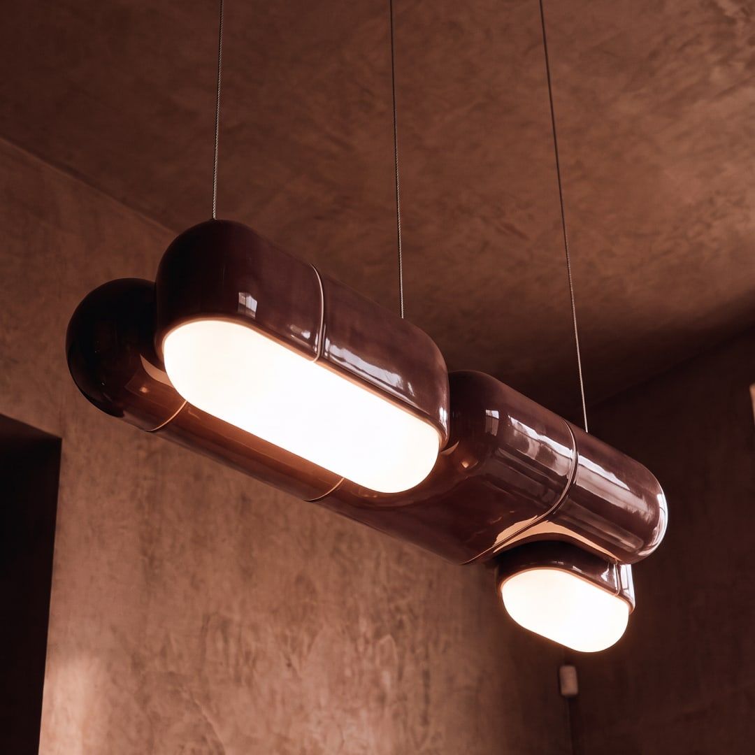 Langwerpige hanglamp in hoogglanzend ossenbloedrood aan dunne kabels, met brede lichtstroken aan de onderkant, voor een roodbruine gestucte wand