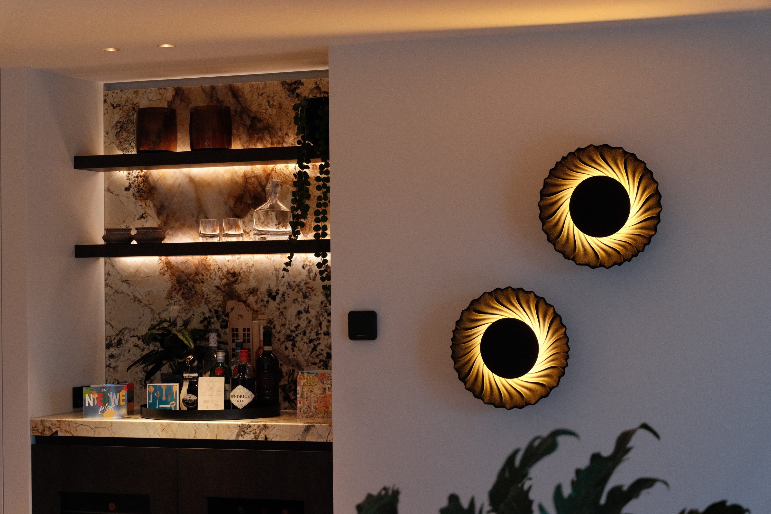 Twee ronde wandlampen met een gouden kraag naast een nis met verlichte planken voor een wand van marmer