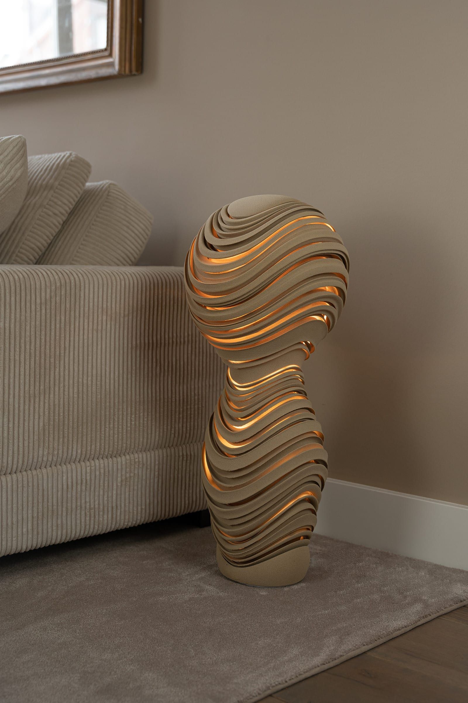 Staande lamp van zandkleurig kwartszand in de vorm van een hoofd op een hals, opgebouwd uit golvende lagen met warm licht ertussen, op een grijs vloerkleed naast een lichte ribfluwelen bank