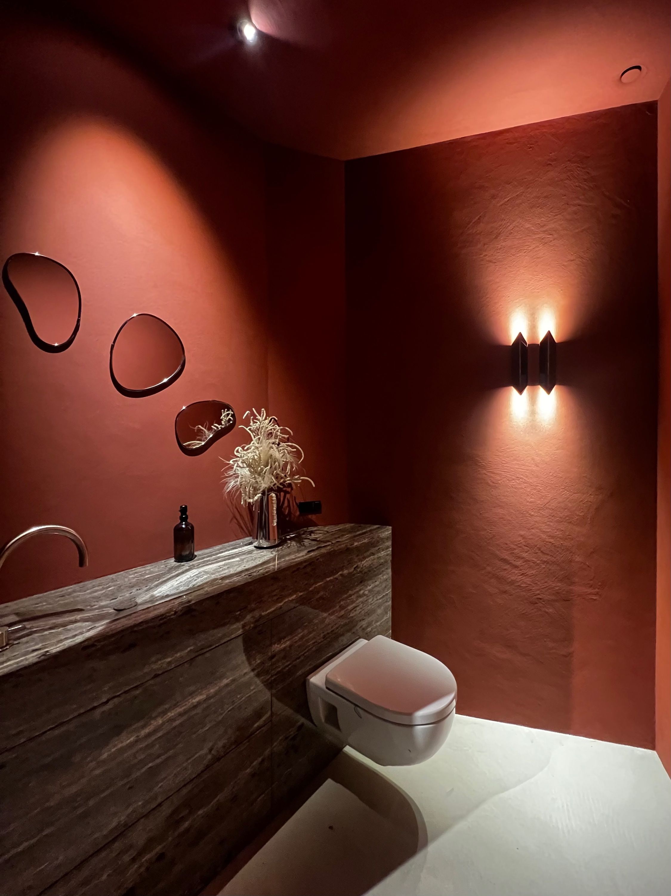 Toilet met terracotta gestucte wanden, een wandlamp die omhoog en omlaag strijkt, drie organische spiegels en een blad van donker natuursteen