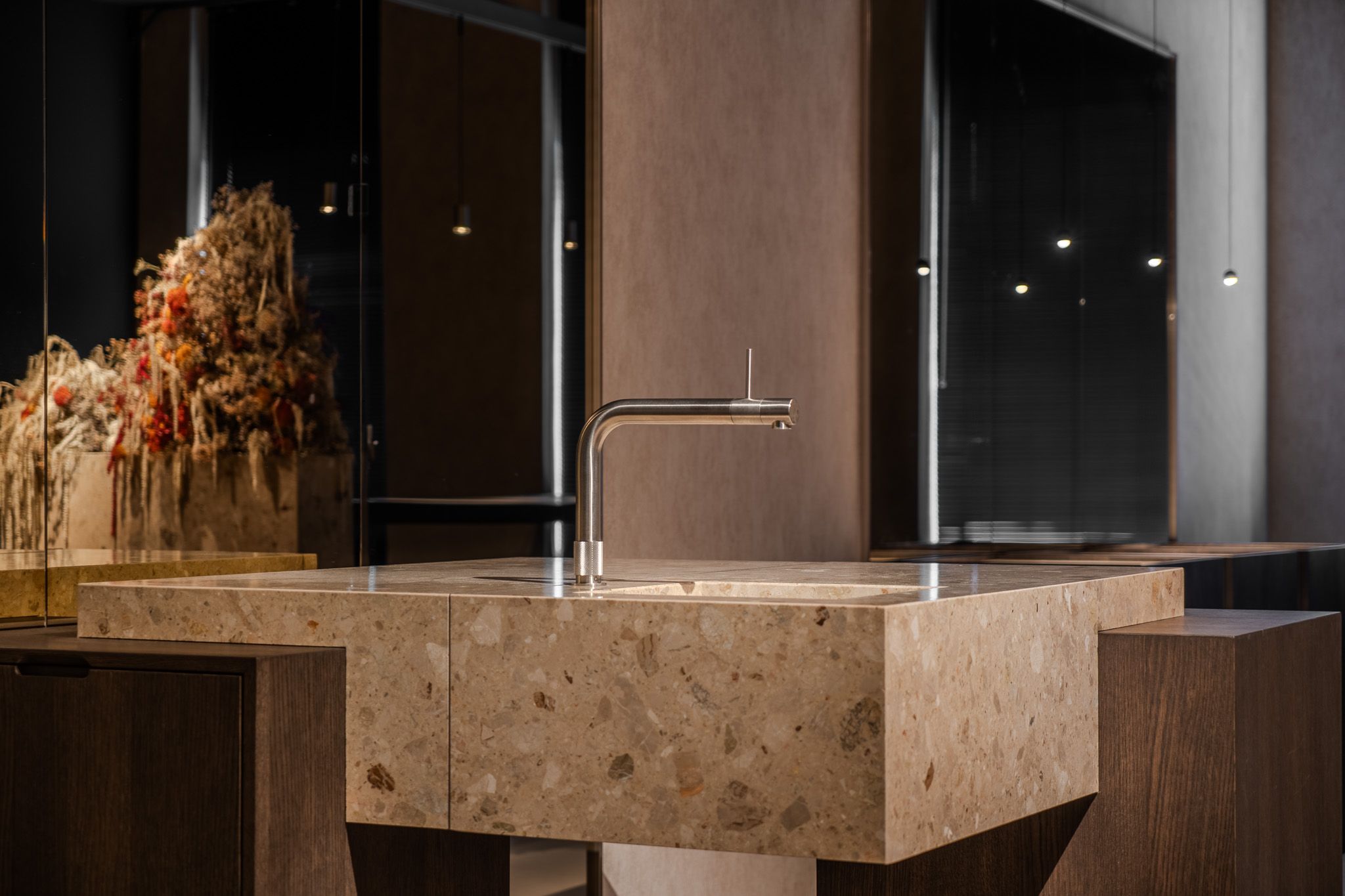 Terrazzo wastafel met een stalen kraan tussen kasten van donker hout, met in de spiegel een bloemstuk van droogbloemen en kleine hanglampjes