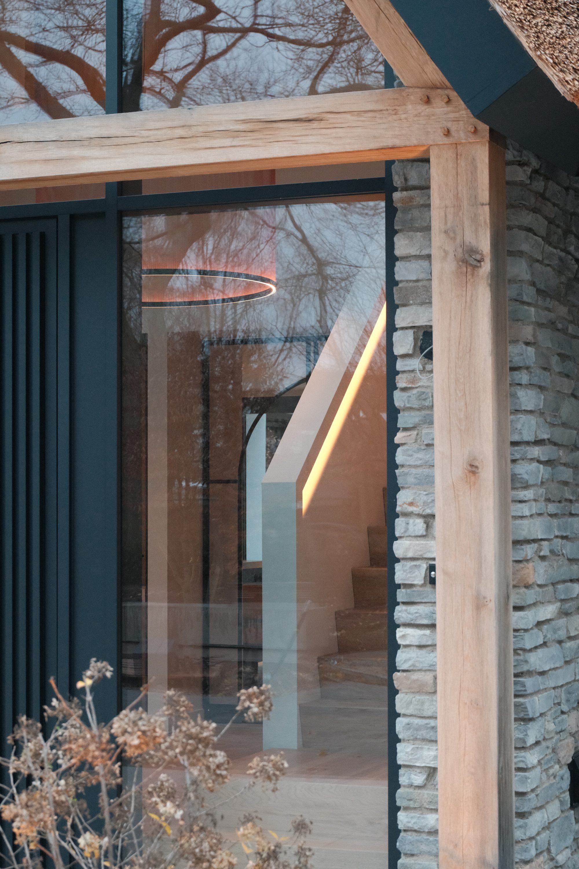 Doorkijk door de glazen gevel naar een trap met een lichtlijn en een ringvormige hanglamp, tussen een eiken balk en een stenen muur
