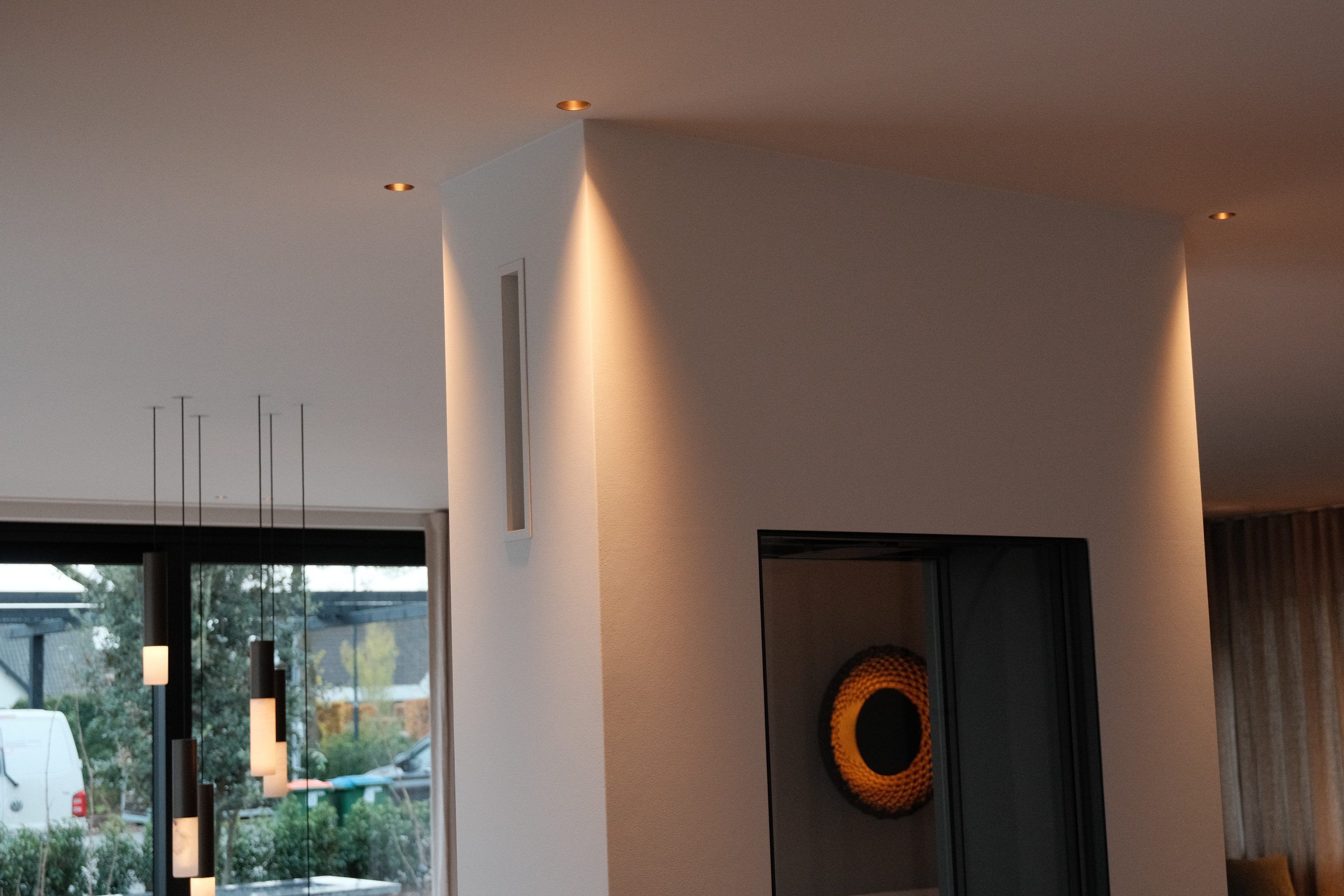 Wand die het licht van inbouwspots opvangt, met een ronde wandlamp in een nis en cilinderhanglampen voor het raam