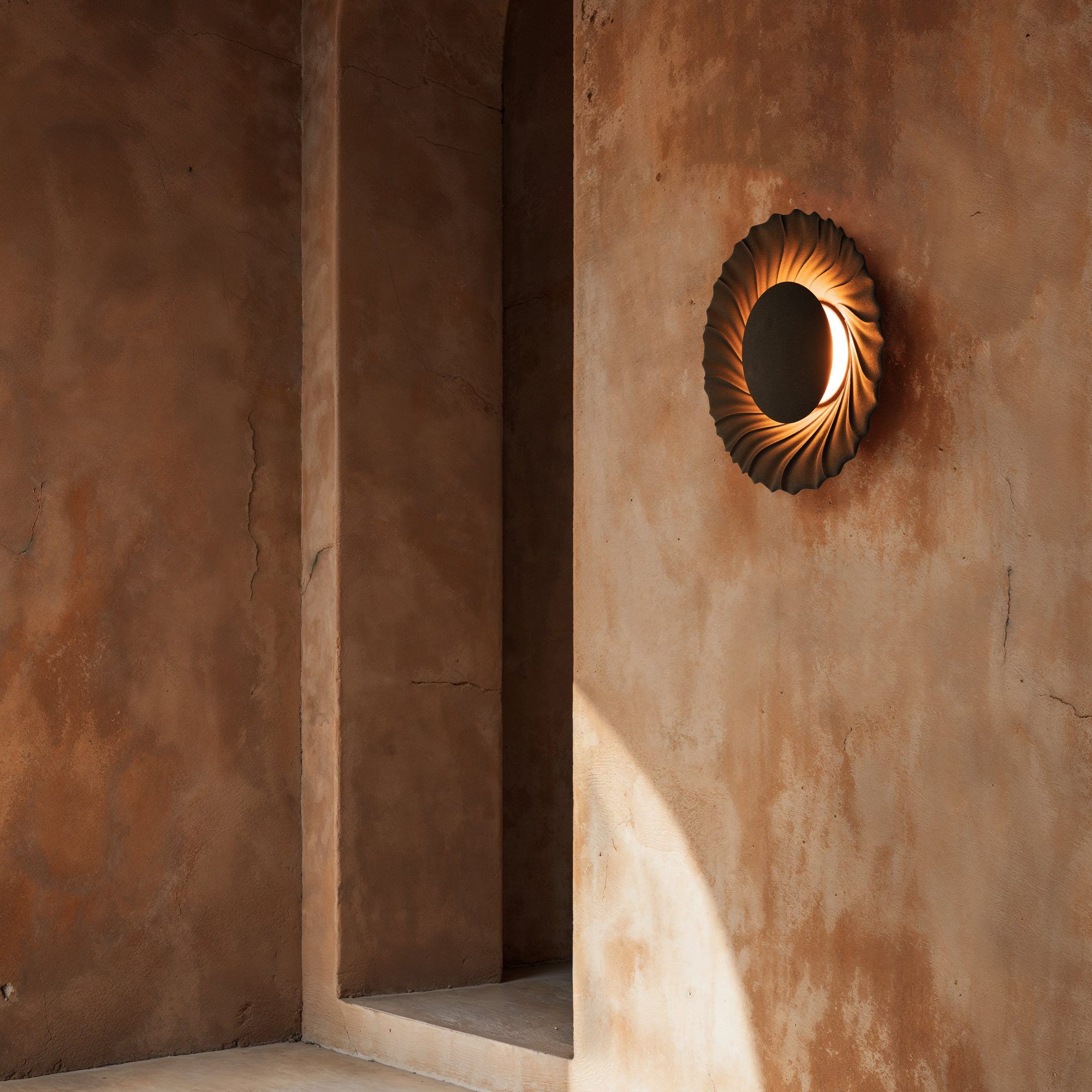 Sun-wandlamp met gedraaide ribben in een lage zon, op een roodbruine gestucte wand naast een boognis