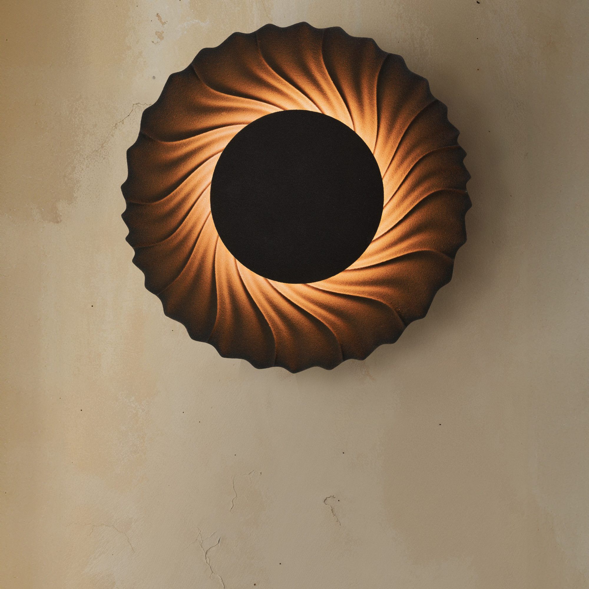 Sun-wandlamp met gedraaide ribben rond een zwarte schijf, het licht valt als een krans over een gestucte wand